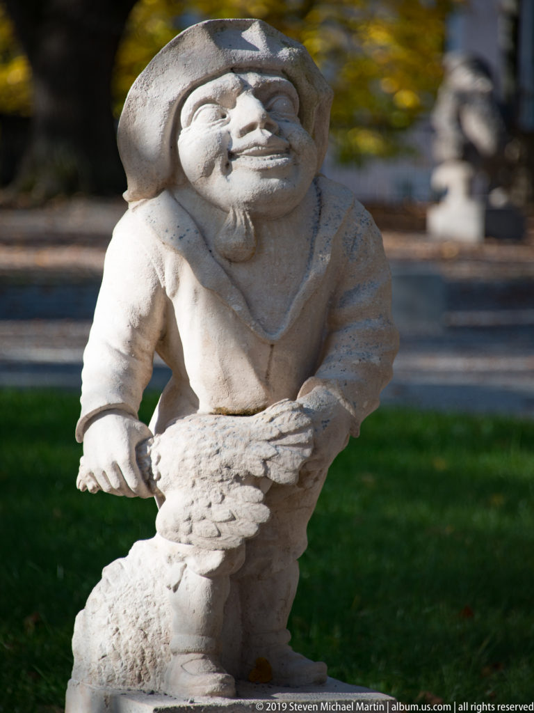 Bearded stone dwarf in a soft cap leaning over a bird-like carved form in front of him.