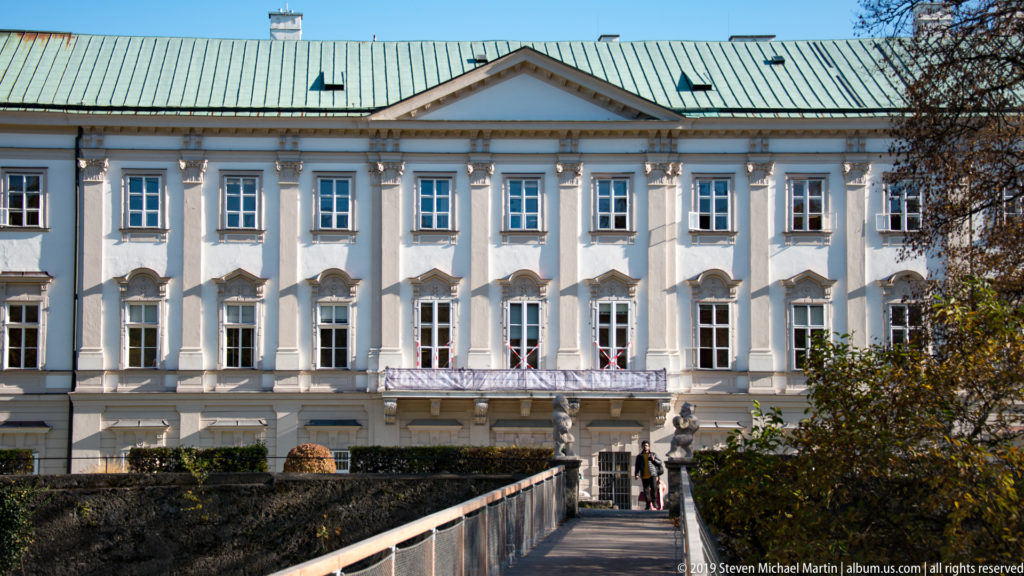 Garden-facing pale Baroque façade of Mirabell Palace with tall windows, central pediment, green roof, hedges, and approach path.