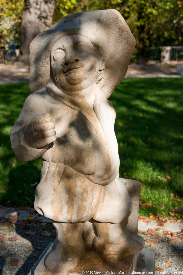 Stone dwarf in a broad hood or cap, smiling with one clenched hand held near the chest.