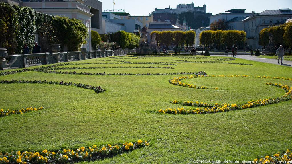 Formal Mirabell Gardens parterre with curving yellow flower beds and clipped trees leading toward Hohensalzburg Fortress in the distance.