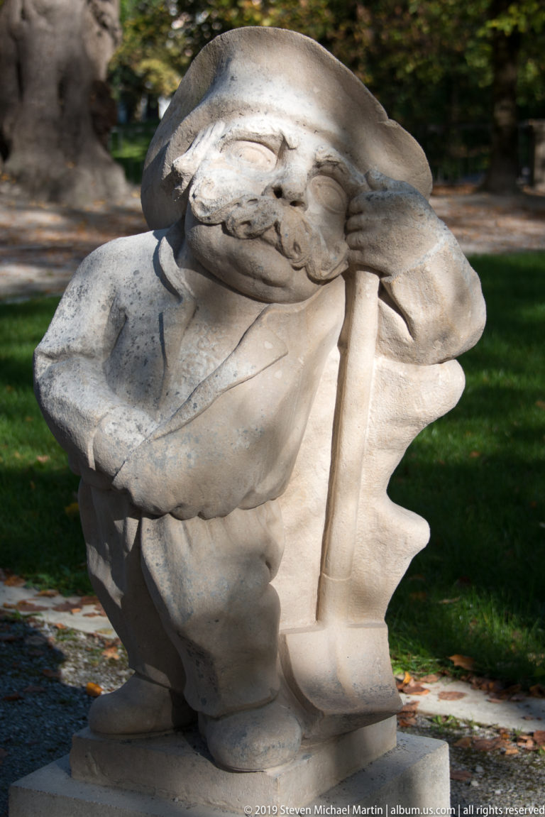 Weary stone dwarf leaning on a long staff with head tilted and one hand pressed against the face.