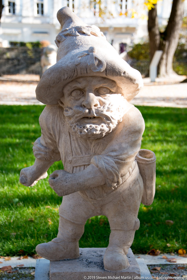 Bearded stone dwarf in a broad hat gesturing with one hand, with a small satchel hanging at his side.