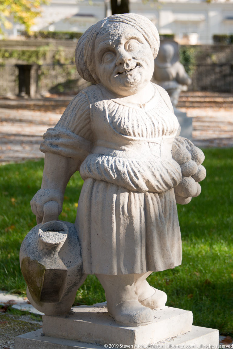Female stone dwarf in a gathered dress standing beside a large jug or vessel on the ground.