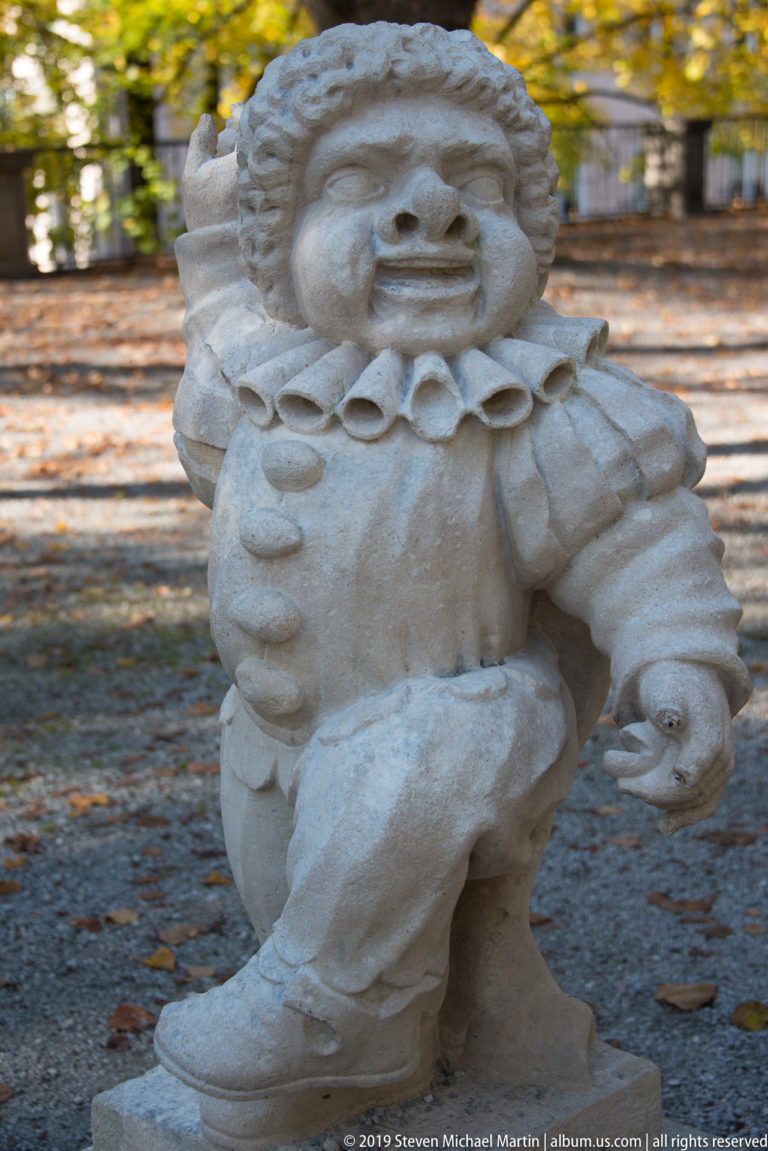 Smiling pale stone dwarf with large ruff collar, puffy sleeves and one hand raised while crouching.