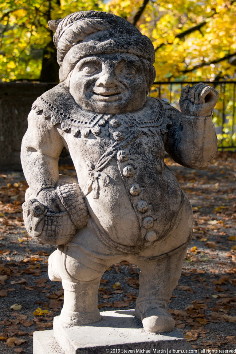 Stout stone dwarf in patterned clothing carrying a cylindrical container and a textured woven object.