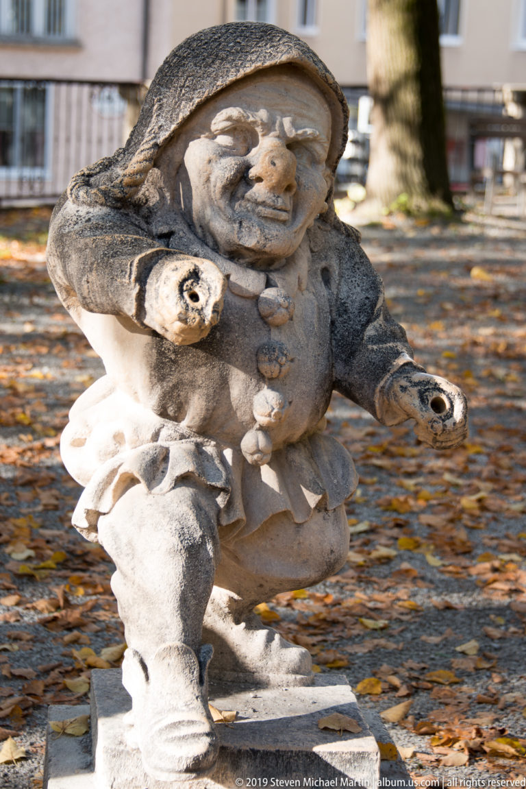 Hooded stone dwarf leaning forward with both hands extended, set against an autumn garden background.