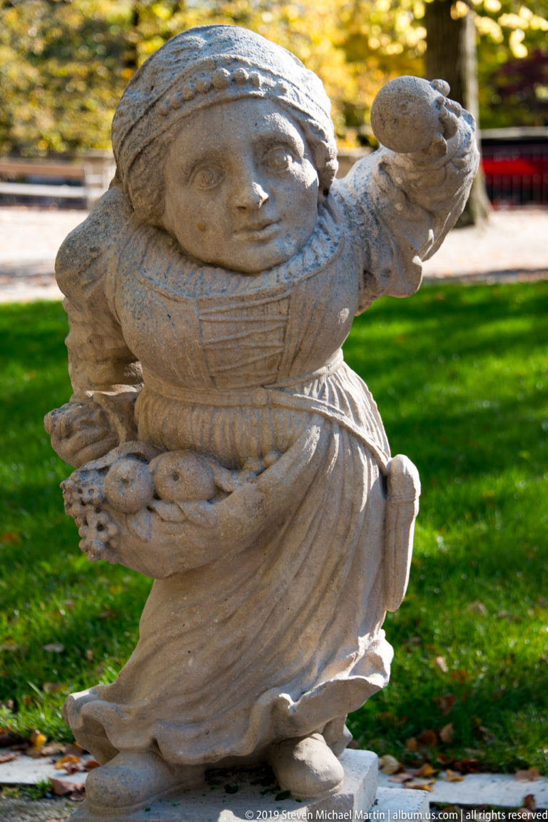 Rounded stone dwarf in patterned costume carrying clustered fruit or produce, with one hand raised.