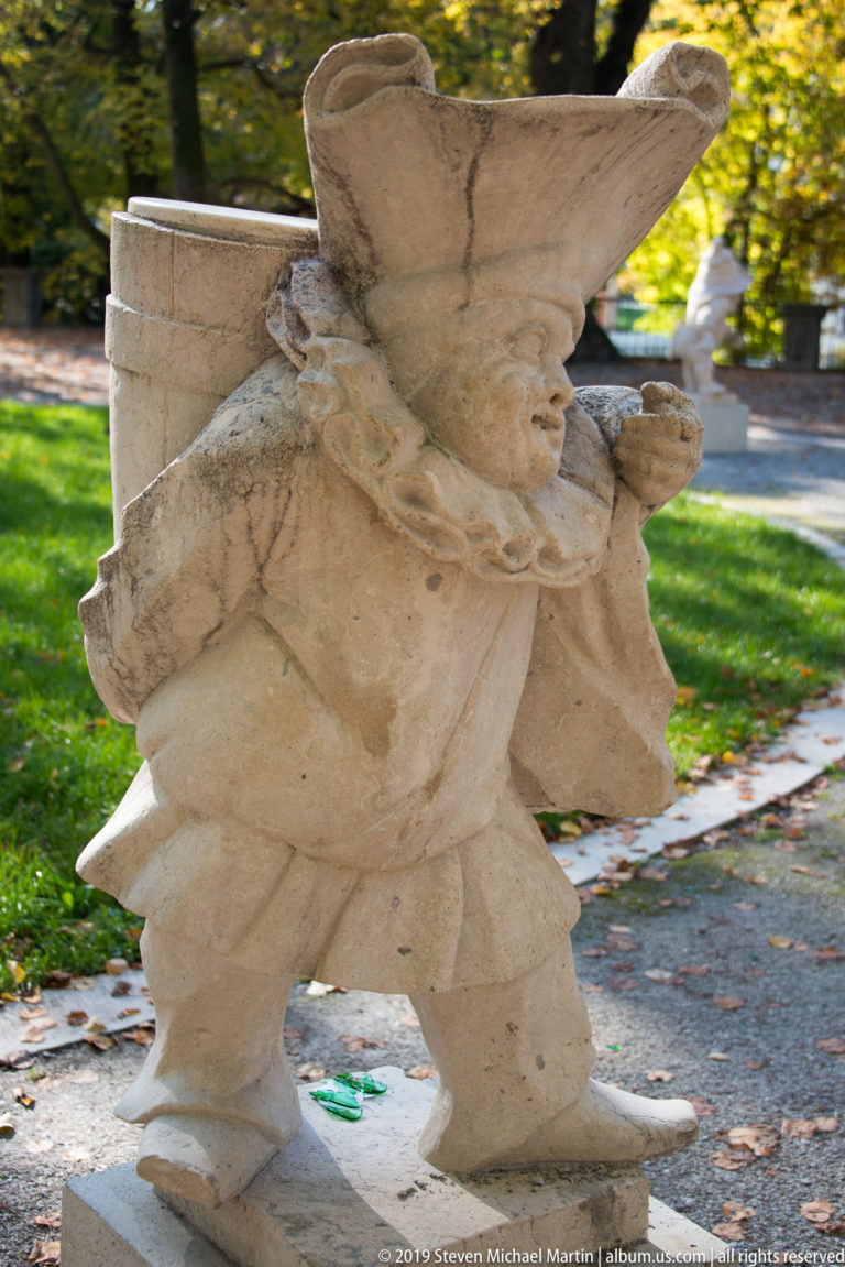 Profile view of a costumed stone dwarf in a broad hat carrying a bulky bundle over one shoulder.
