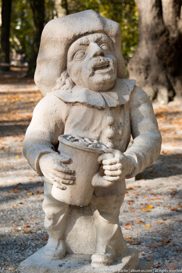 Smiling stone dwarf in broad hat and ruff collar holding a large vessel at waist height.