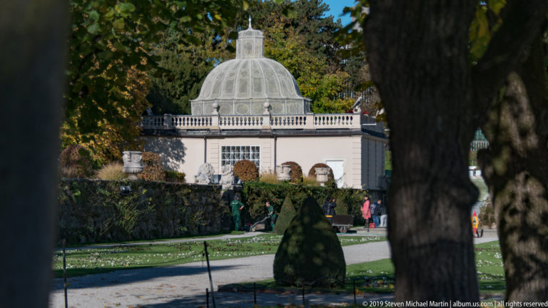 Small pale garden building with ribbed dome and balustrade seen through trees and clipped shrubs in Mirabell Gardens.