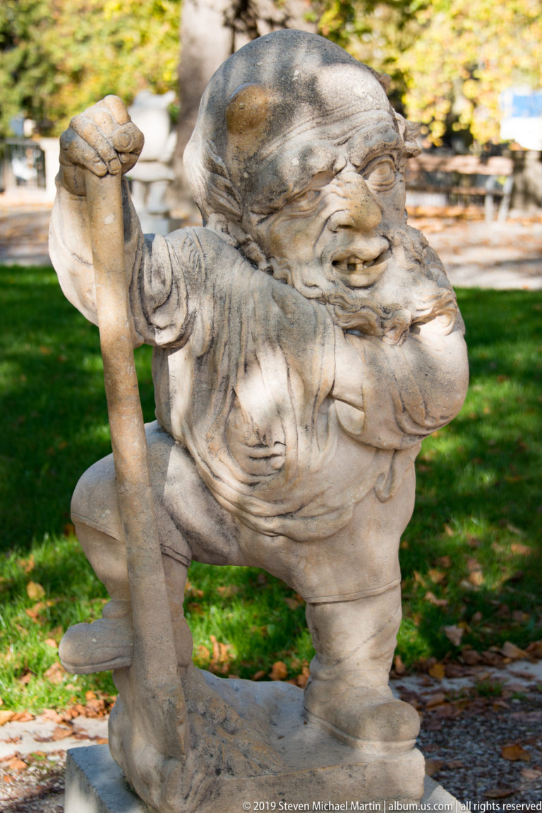 Stooped bearded stone dwarf leaning on a long staff in a garden scattered with autumn leaves.