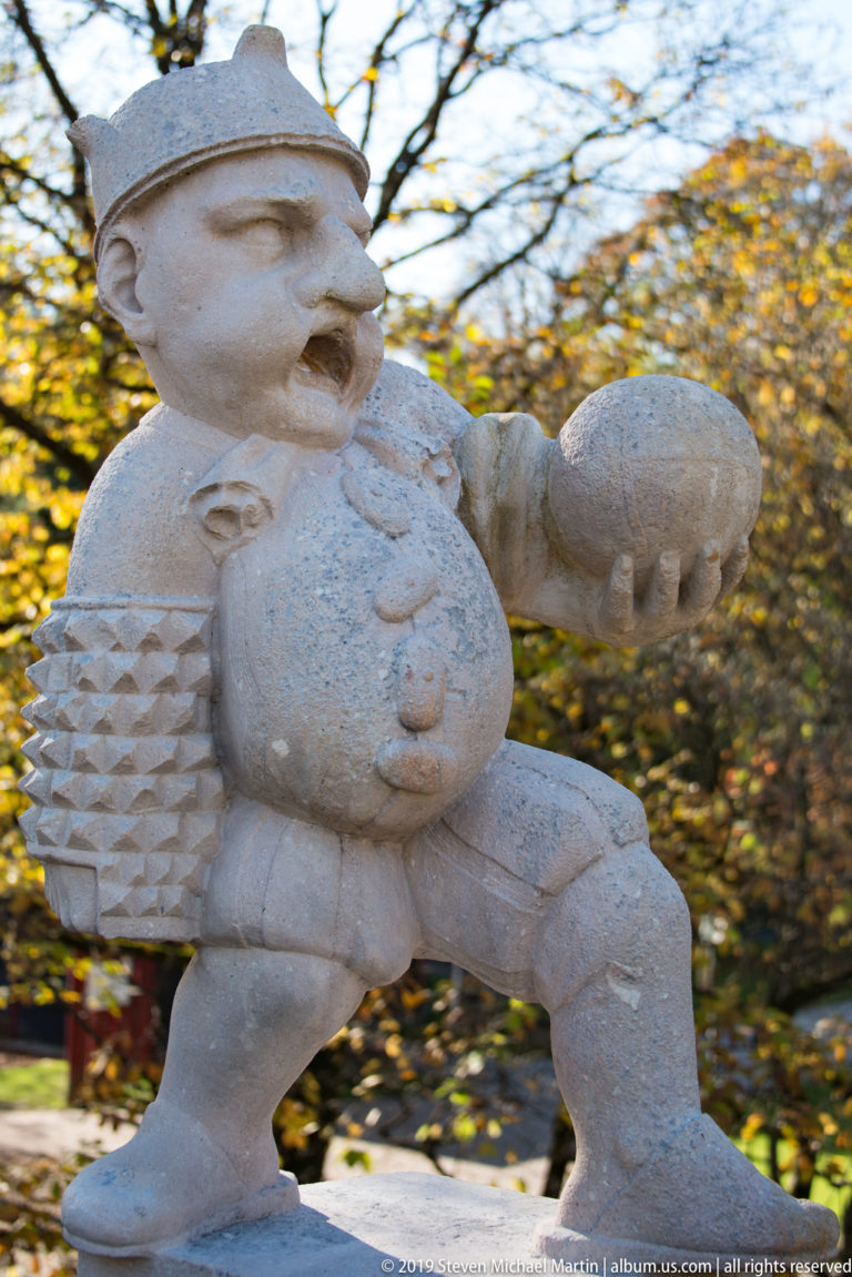 Caricatured stone dwarf with prominent belly and textured sleeves, striding while holding a large rounded object.