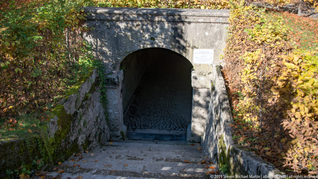 Low stone archway leading into a shadowed cobbled passage beneath a leaf-covered garden embankment.