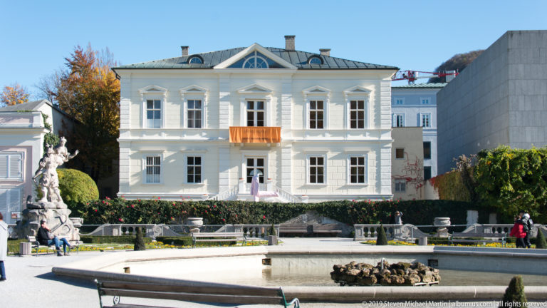 Small symmetrical white garden building with tall windows and central pediment beyond a broad stone basin and statues.