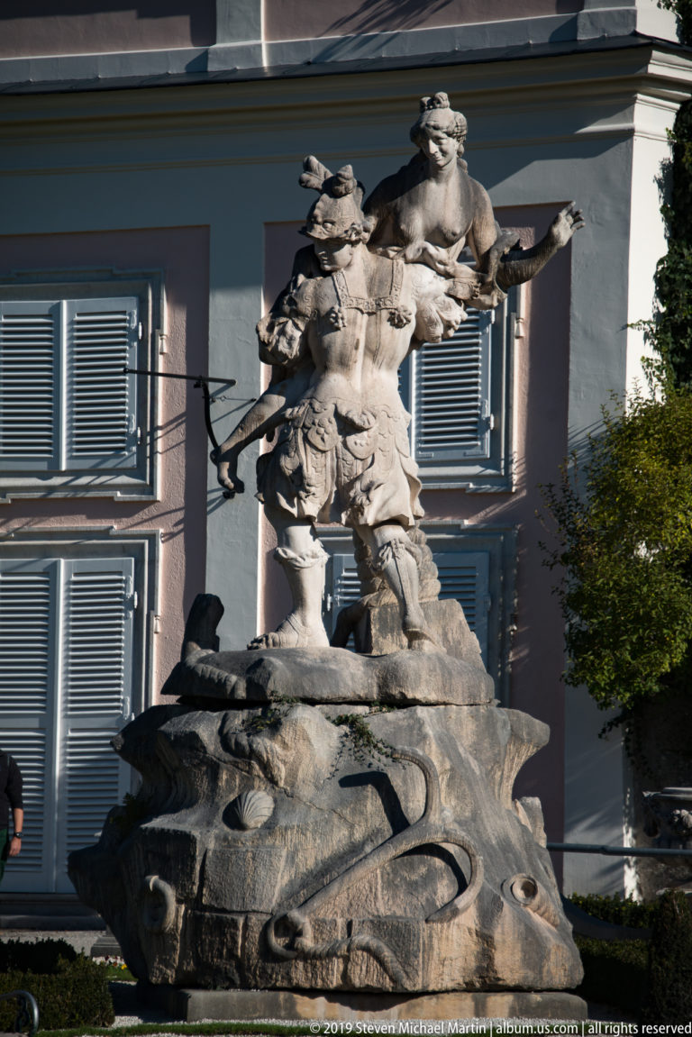 Complex stone sculpture with human figures and a large animal form on a carved base beside a palace façade.