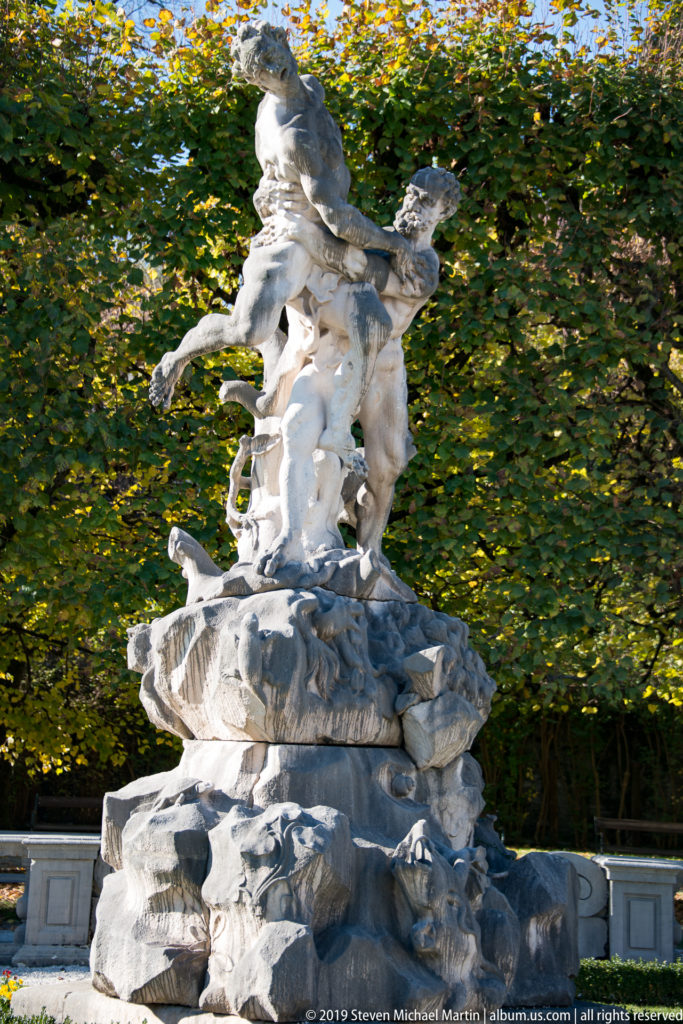White Baroque mythological sculptural group on a rocky fountain base amid green foliage in Mirabell Gardens.