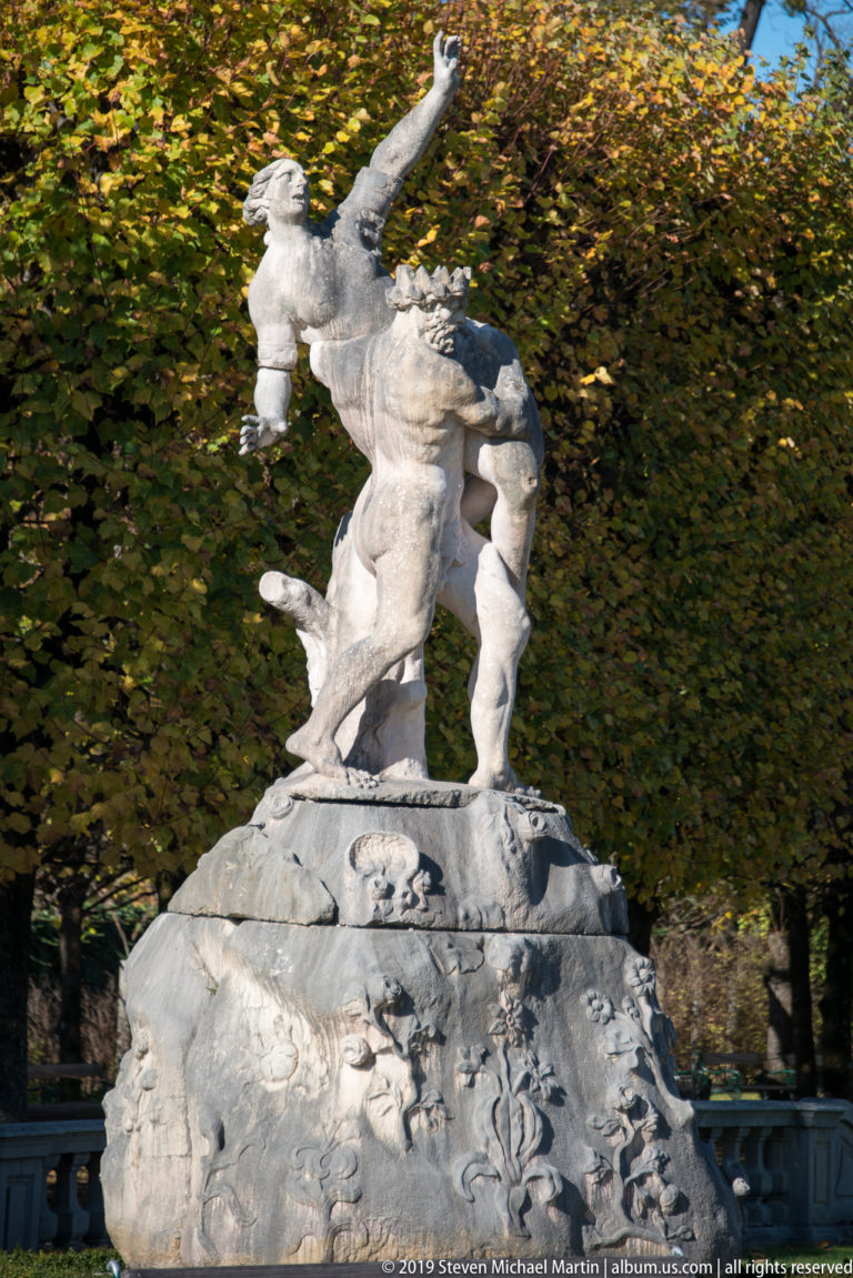 Muscular pale stone garden statue with one arm raised, standing on a carved rockwork base before autumn trees.