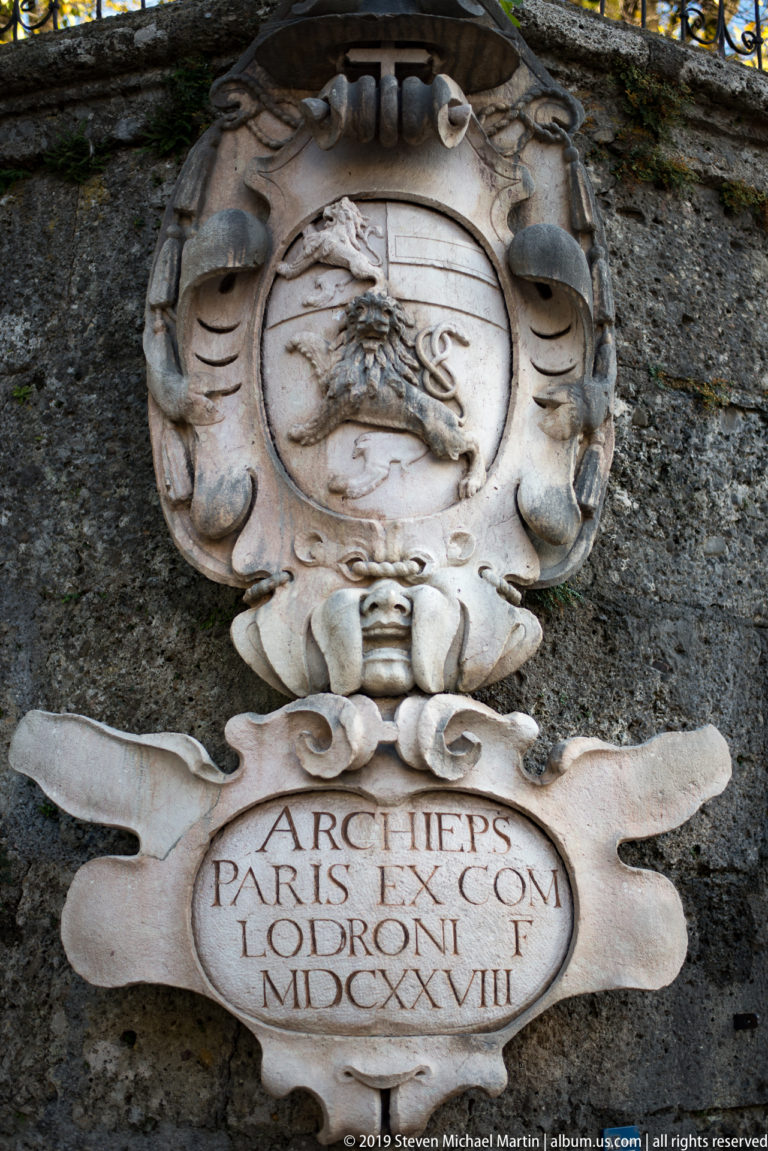 Carved stone coat of arms above a draped Latin-inscribed tablet mounted on a dark masonry wall.