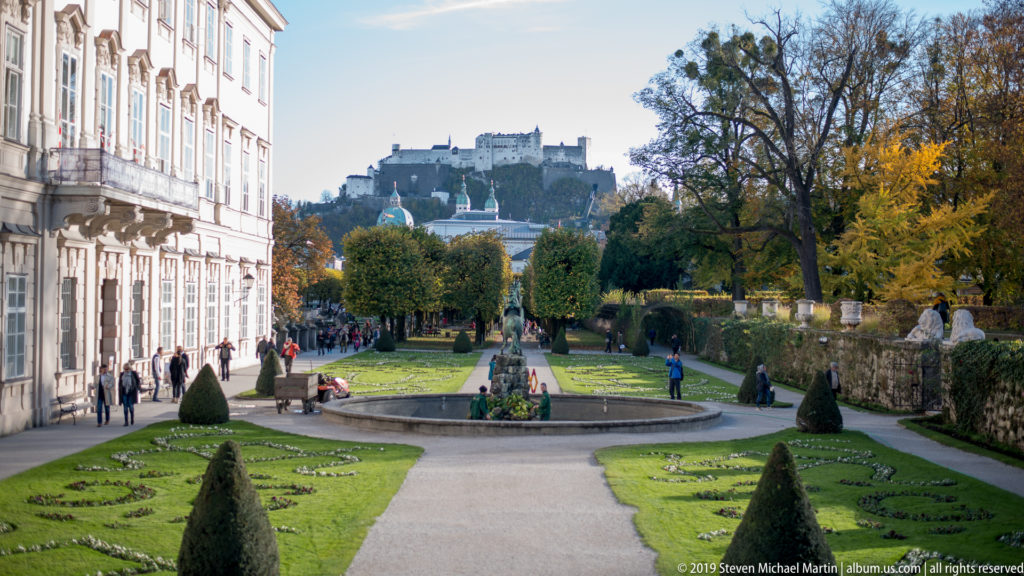 Formal Mirabell Gardens with clipped lawns and fountain leading toward Hohensalzburg Fortress on a hill beneath a blue sky.