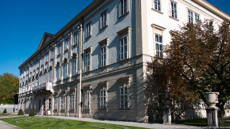 Long pale side façade of Mirabell Palace with rows of tall windows beside lawn, clipped plantings, and garden ornament.