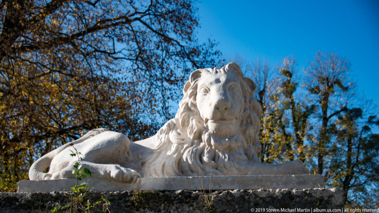 Frontal view of a reclining pale stone lion with full mane and one forepaw extended on a garden plinth.