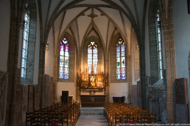 Gothic chapel interior with rib vaulting, three stained-glass windows, rows of chairs, and a winged altarpiece at the far wall.