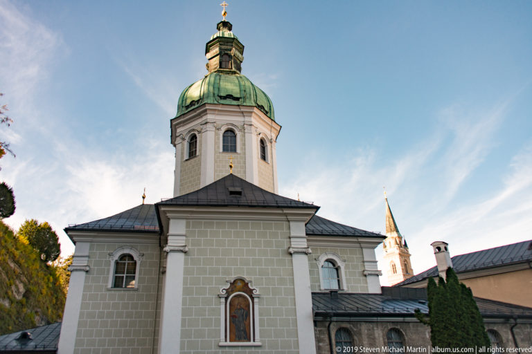 Pale domed chapel with arched windows and green copper roof in St. Peter's Cemetery, with a distant church spire.