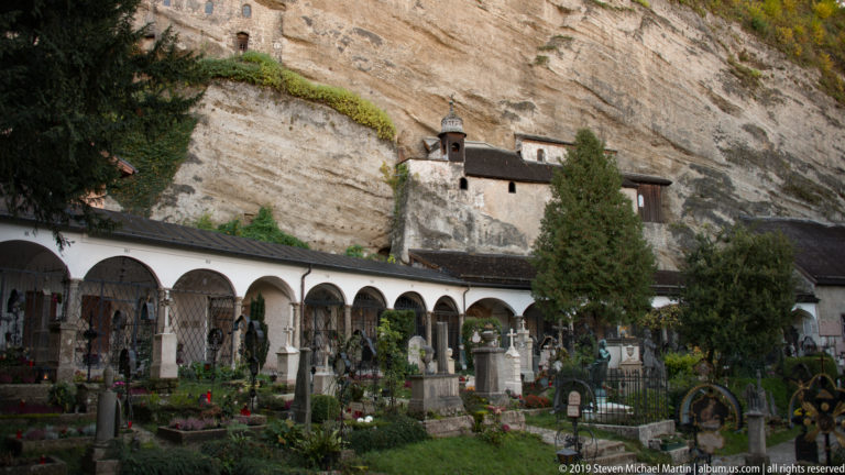 Historic cemetery with planted graves and arcades beneath a sheer rock cliff, with a small chapel-like structure built against the rock.