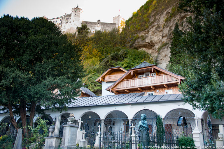 White cemetery arcade with iron grave enclosures and memorials below timber roofs, cliff, and Hohensalzburg Fortress.