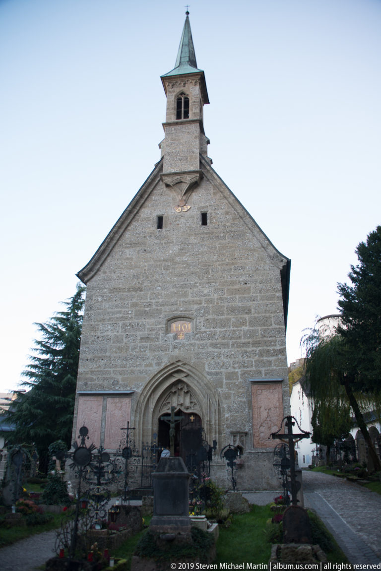 Gabled stone chapel with pointed portal and small bell turret amid graves and trees in St. Peter's Cemetery, Salzburg.