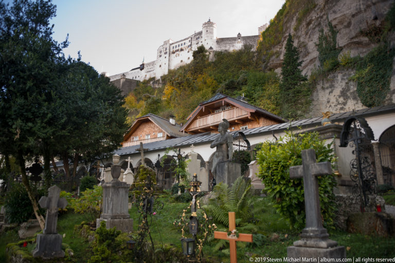 Historic cemetery with crosses and planted graves below a cliff and Hohensalzburg Fortress in Salzburg.