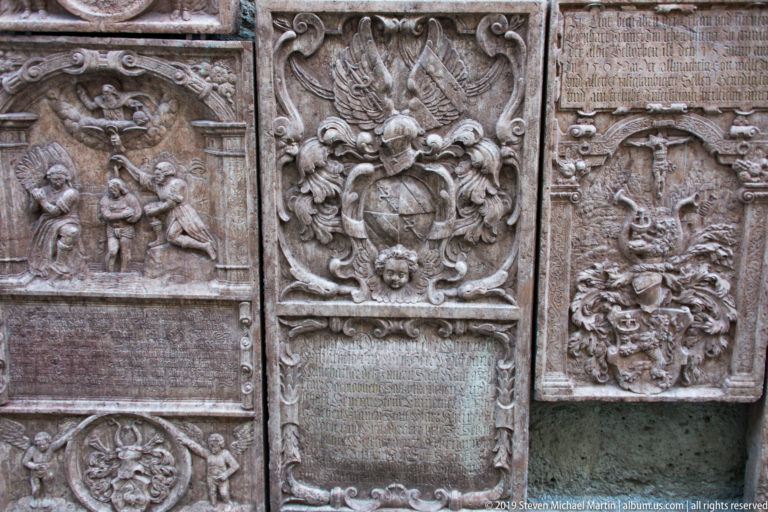 Closely grouped historic grave slabs with carved inscriptions, heraldic shields, cherubs, and Baroque relief ornament.