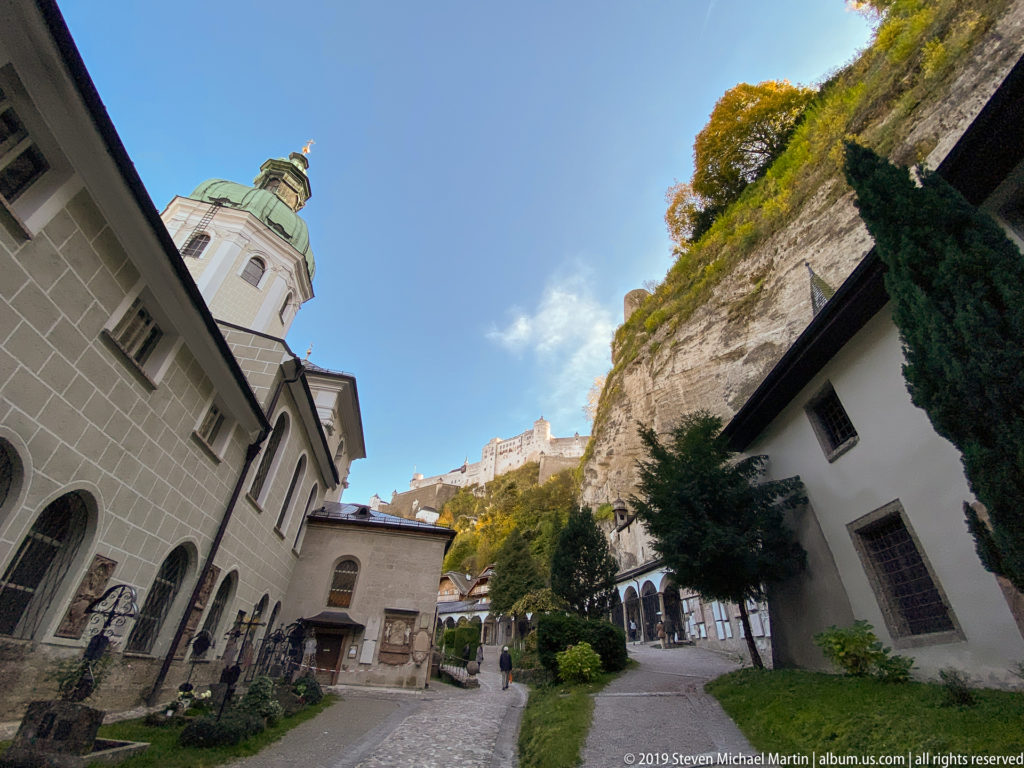 St. Peter’s Abbey buildings and cemetery structures below a steep cliff, with Hohensalzburg Fortress visible high on the ridge.