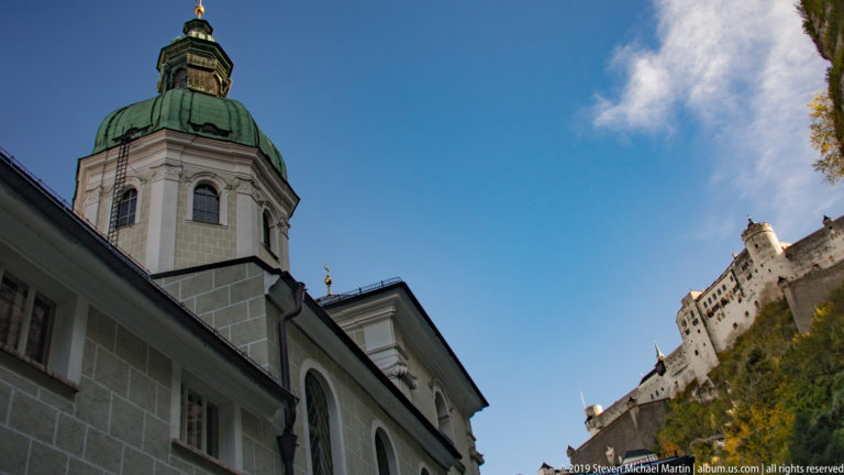 Pale chapel with green copper dome and tall arched windows below Hohensalzburg Fortress on the rocky hillside.