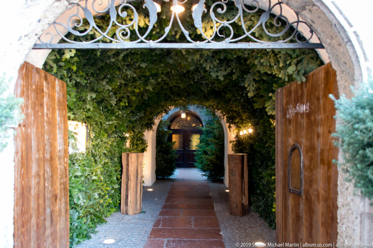 Open wooden gates leading along a red-paved, ivy-lined walkway through an arched garden passage.