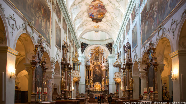 Long view down the abbey church nave toward the ornate high altar, with side arches, paintings, sculptures, and patterned floor.