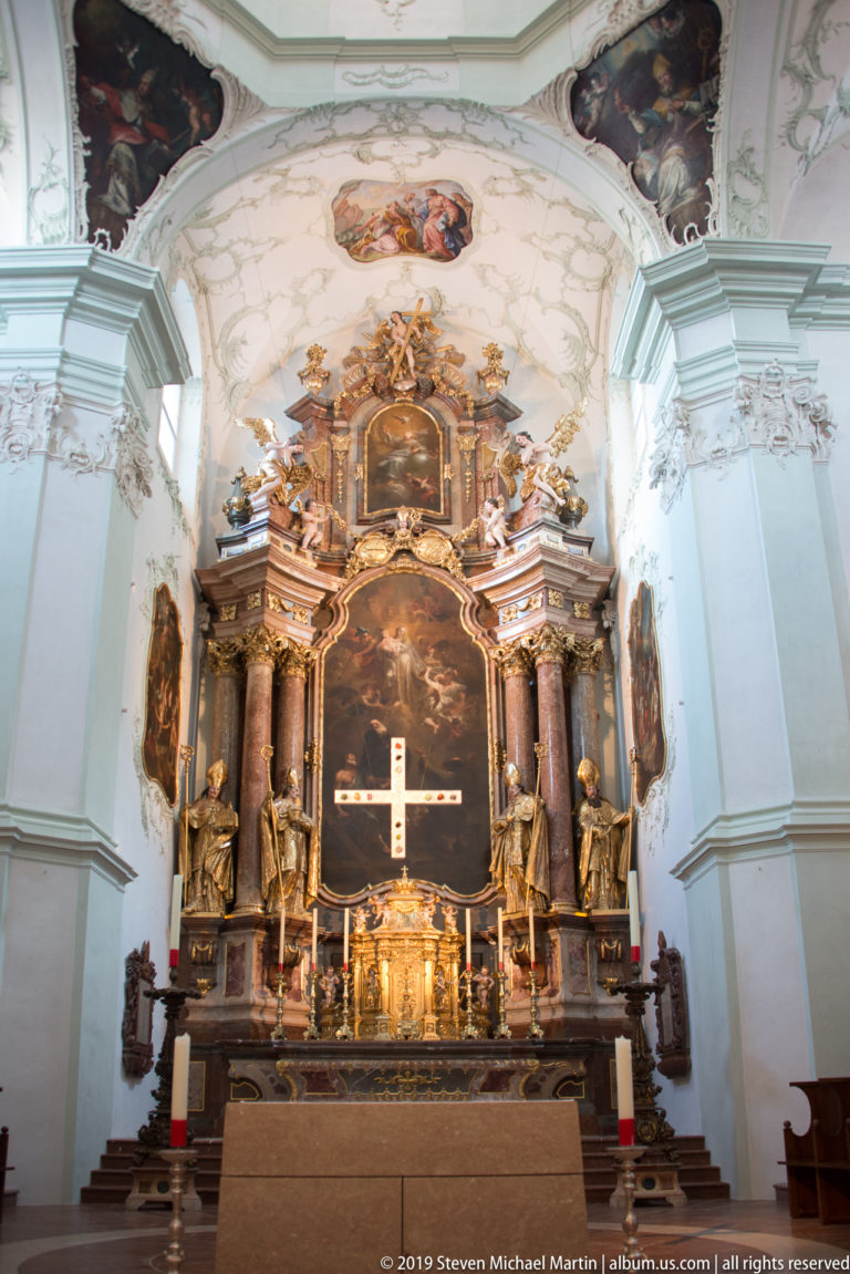 Baroque high altar with dark columns, gilded figures, central altarpiece, cross, and candlesticks beneath a painted ceiling.