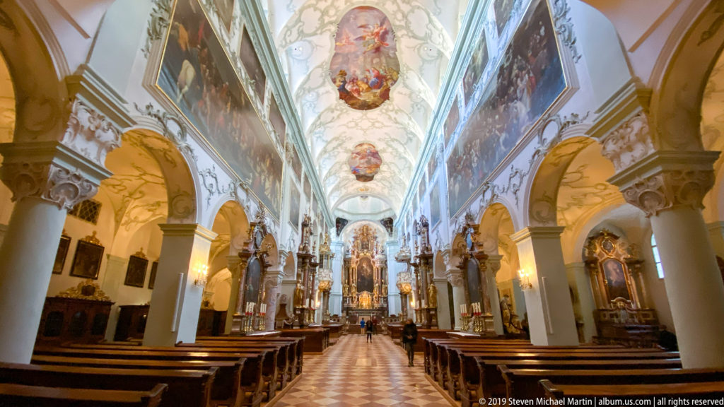 Centered nave view toward the sanctuary, framed by white piers, gilded capitals, ceiling paintings, side altars, and wooden pews.