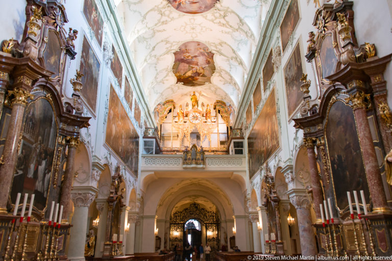 Decorated abbey church nave viewed upward toward a gallery, painted vault, side altars, and large wall paintings.