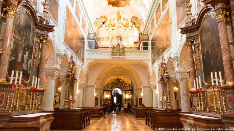 Long decorated church nave with side altars, paintings, gilded furnishings, vaulted ceiling, and visitors near a central arch.