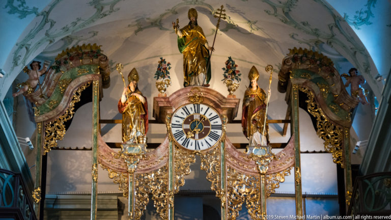 Ornate clock with three gilded saint figures, carved scrollwork, and curved decorative framing in the abbey interior.