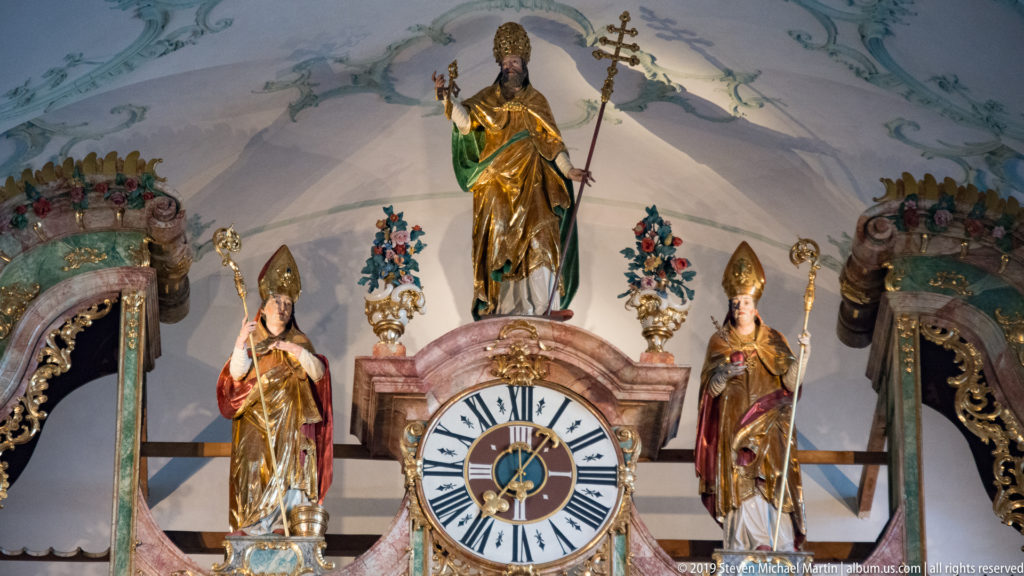 Large clock framed by gilded bishop saints and ornate Baroque decoration inside St. Peter's Abbey.
