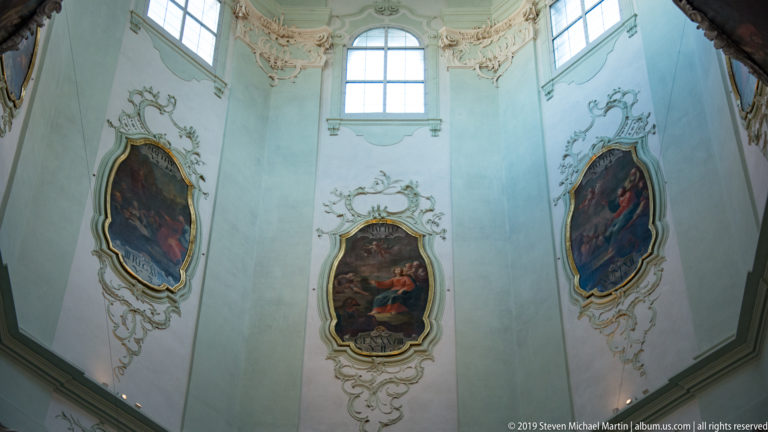 Three framed religious paintings set into a pale wall with ornate Rococo stucco beneath a tall arched window.