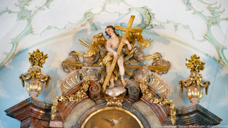 Polychrome figure of Christ carrying the cross above an ornate gilded structure with angels and Rococo stucco.