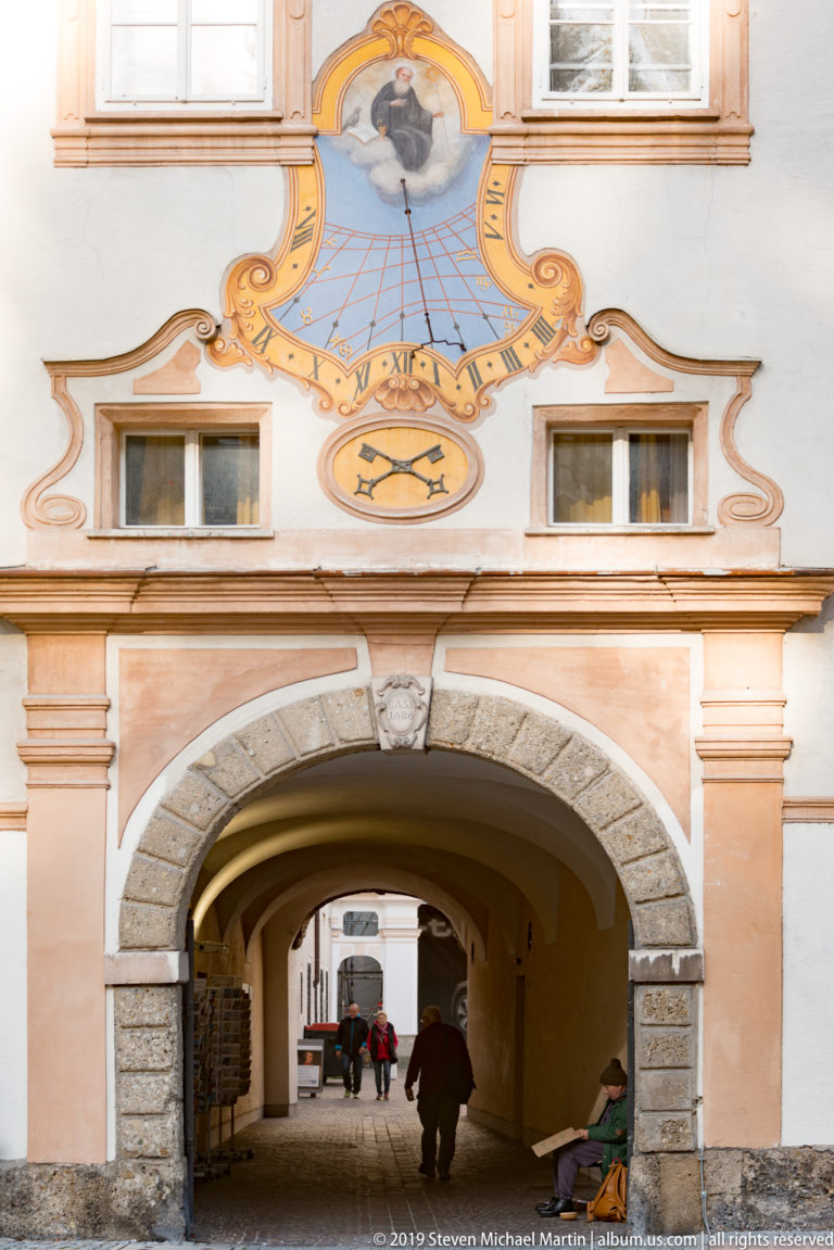 Large arched abbey passage with rusticated stone surround beneath an ornate painted sundial and windows.