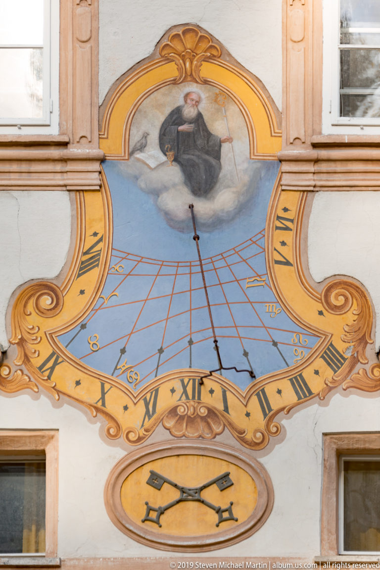 Decorative wall sundial with Roman numerals, painted religious figure, scrolling ornament, and crossed keys on the abbey façade.