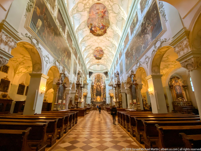 Central view down the nave of St. Peter’s Abbey Church with pews, arcades, frescoed ceiling, side altars, and high altar.