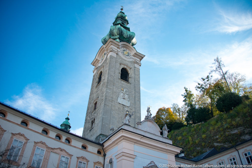 Pale stone abbey bell tower with green onion dome above pink-and-white courtyard buildings under a blue sky.