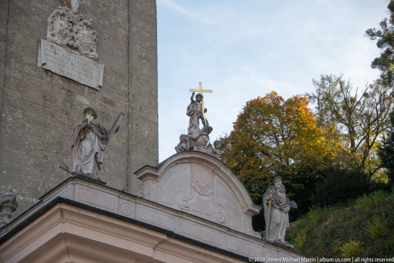 Stone religious statues arranged along an abbey roofline beside a tall masonry wall and wooded hillside.