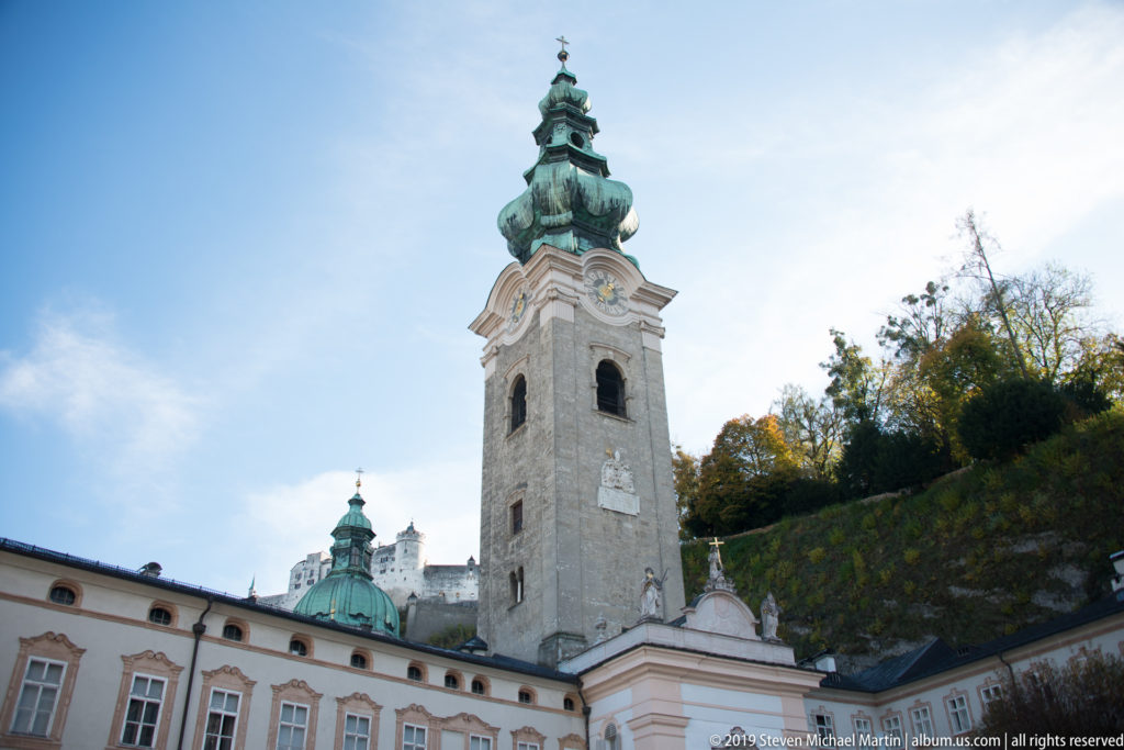 Tall stone church tower with clock, arched belfry openings, and green onion dome above the abbey courtyard in Salzburg.